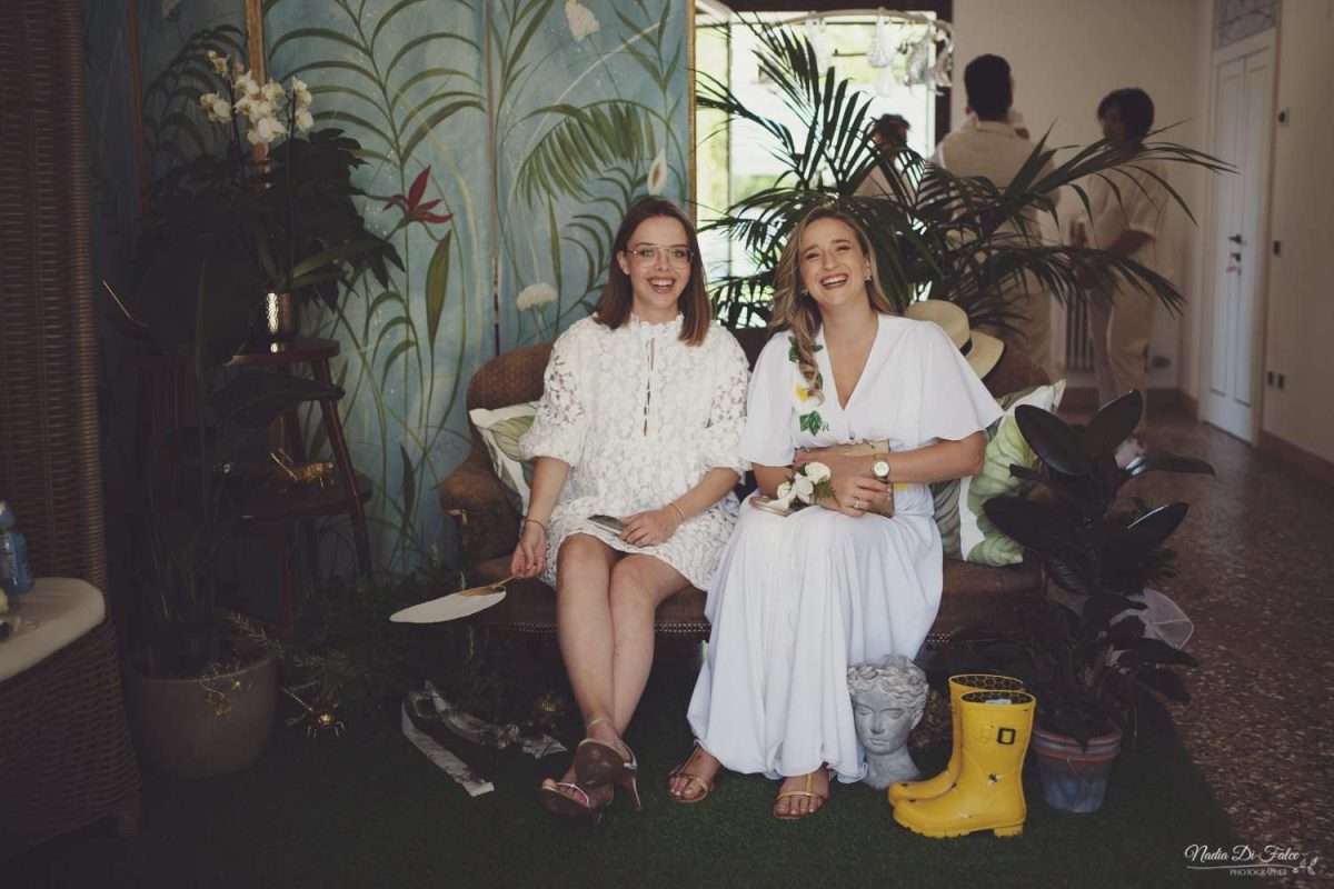 Damigelle sorridenti di Maria Mayer sedute sul divanetto di casa, in abito bianco